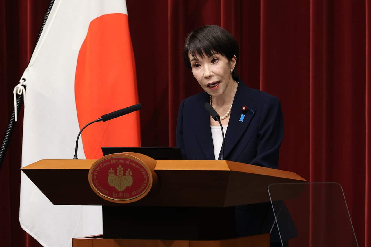 A woman speaking at a podium, flanked by a Japanese flag.