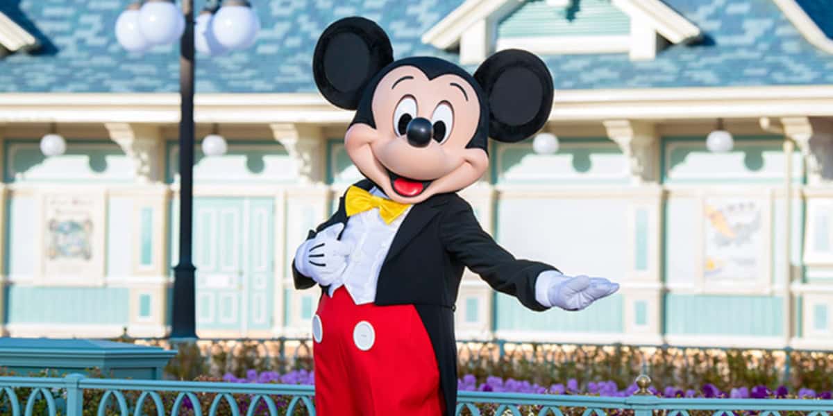 Mickey Mouse, wearing his iconic red trousers, yellow bow tie, and white gloves, poses with an inviting gesture in front of a pastel-colored, Victorian-style building. The setting appears bright and cheerful, with a clear day and decorative lighting at this Tokyo Disney Park.