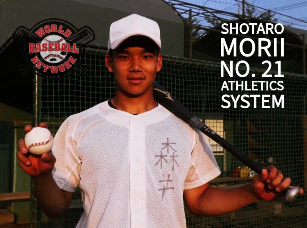Young baseball player in a white uniform holds a baseball and rests a bat on his shoulder in front of a batting cage.