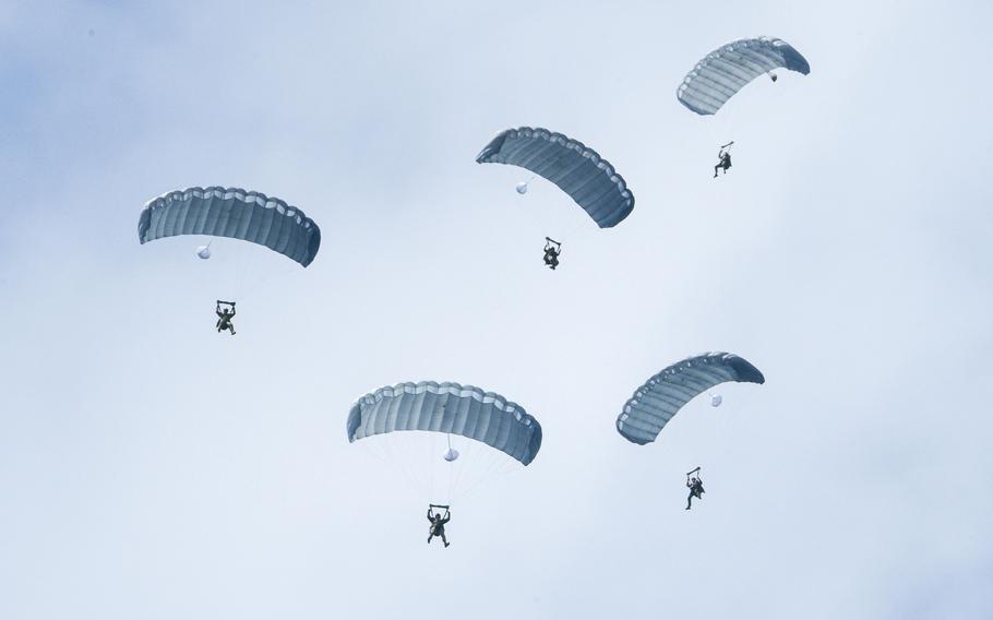 Five military service members with parachutes, seen from below, descend to the ground.