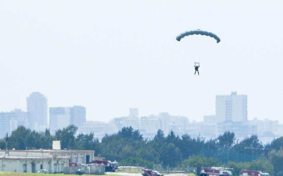 A military service member with a parachute, seen from a distance, descends to the ground, with a city skyline in the background.