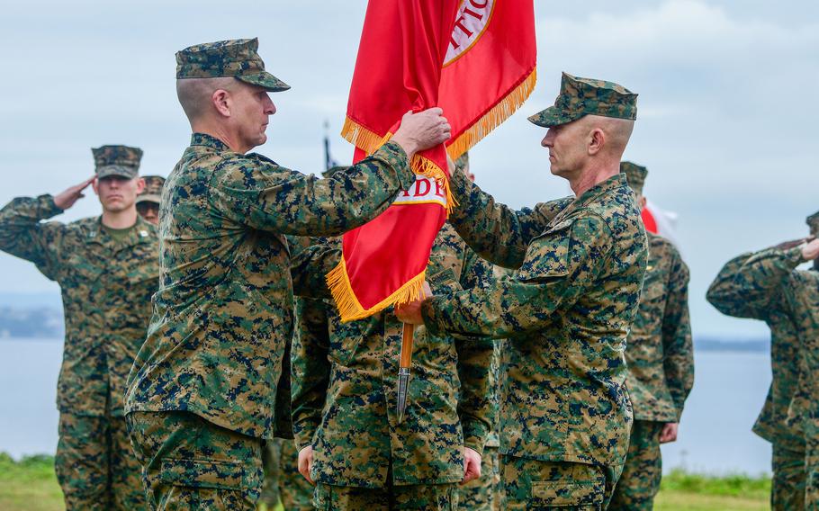 Service members salute in the background as the Marine Corps flag is handed off during an outdoor change-of-command ceremony.