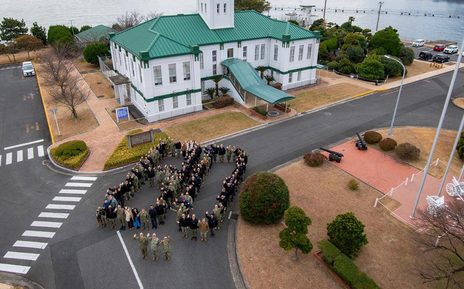 A large group of people, seen from the air, gather to form the number 80 on a street in front of a white and green building.