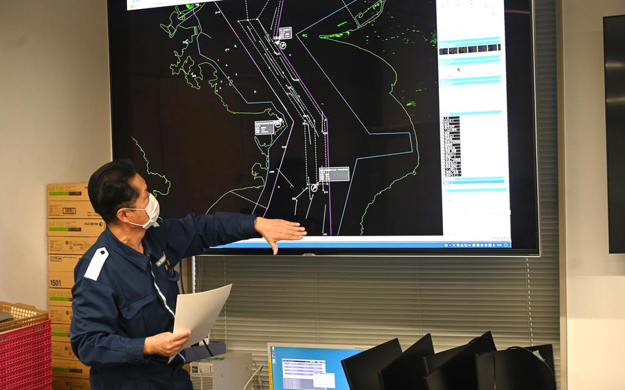 A man in a blue uniform points at a a large monitor while gesturing.