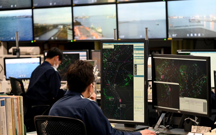 A close view of an operations desk where a person is working at multiple monitors displaying detailed vessel‑tracking maps and navigational data. Papers, equipment, and consoles are arranged across the workstation.