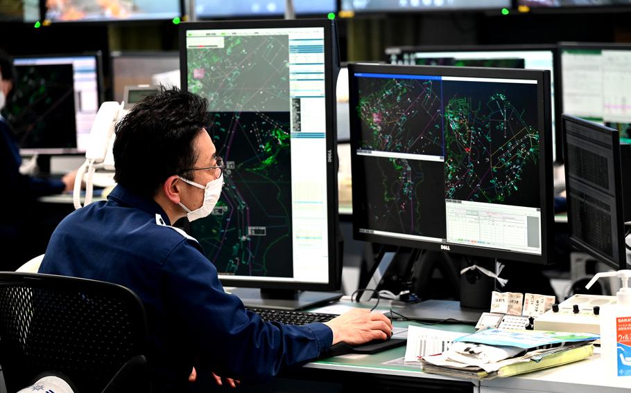 A close view of an operations desk where a man in a dark blue uniform is working at multiple monitors displaying detailed vessel‑tracking maps and navigational data. Papers, equipment, and consoles are arranged across his workstation.
