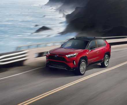 Dynamic front 3/4 shot of a red 2024 Toyota RAV4 Prime XSE driving on a road running beside the ocean.