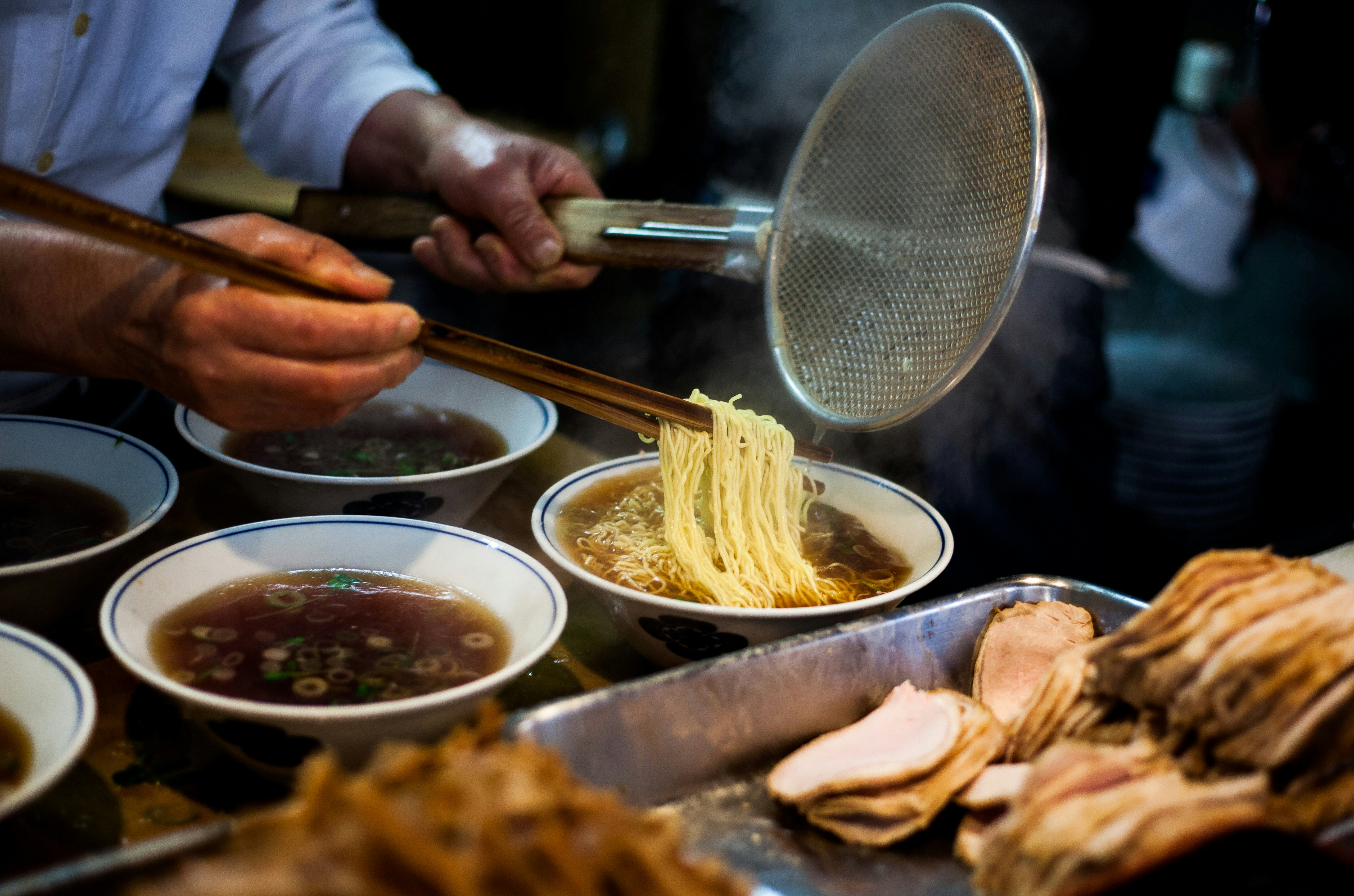 Hands preparing steaming ramen (soba) while holding long chopsticks and metal strainer. Several bowls of broth and pork are in the foreground.