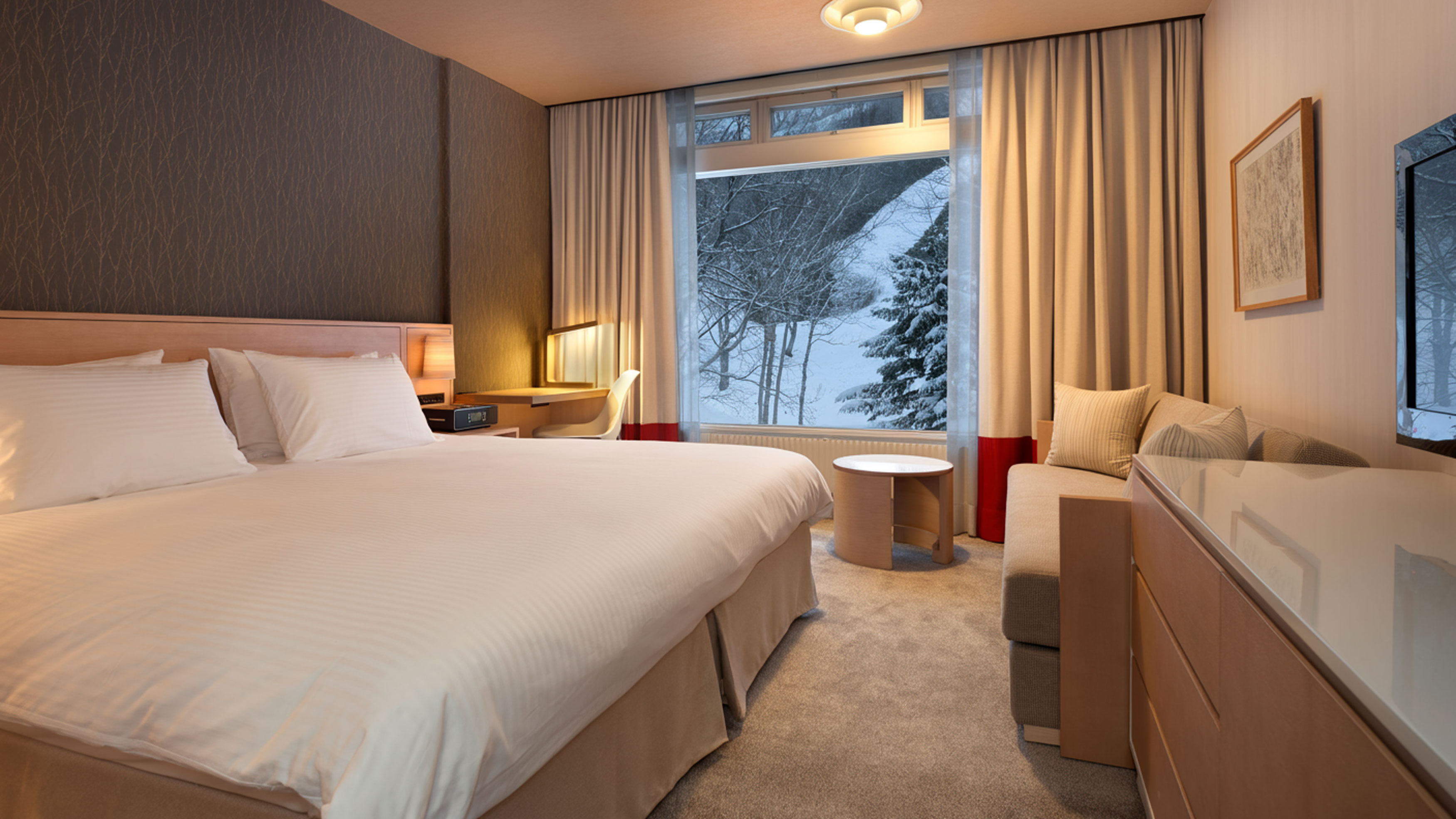 a room with king bed, couch, desk, tv, and large window looking at snowy landscape