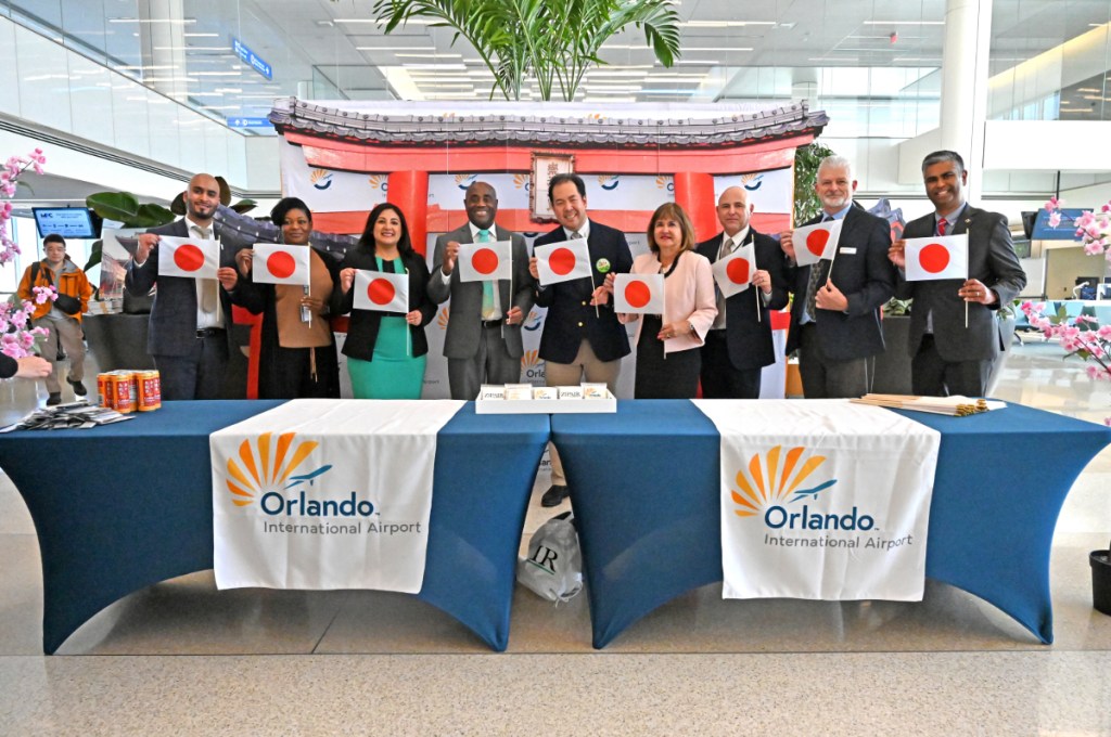 A group of people holding papers with the Japanese flag while standing behind two blue tables with a table covering that reads "Orlando International Airport"