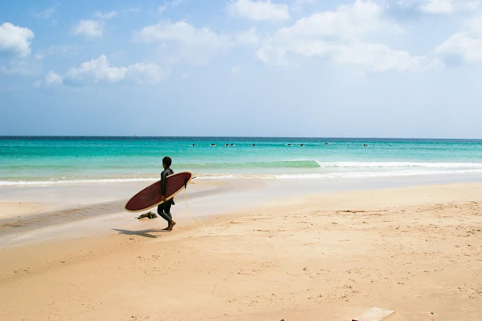A wide, soft-sanded beach with shallow waves crashing in the distance as a young surfer walks by carrying his surf board.