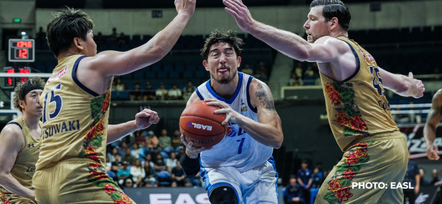 A basketball player in a blue jersey attempts to drive to the basket while being guarded by two defenders in patterned jerseys during a competitive game.