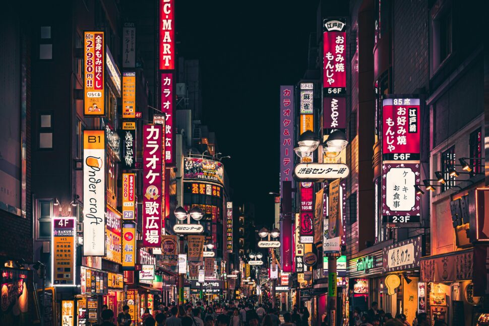 A busy street in Tokyo at night filled with brightly lit neon signs in Japanese, colorful storefronts, and crowds of people walking beneath the glow of red, yellow, and purple billboards.