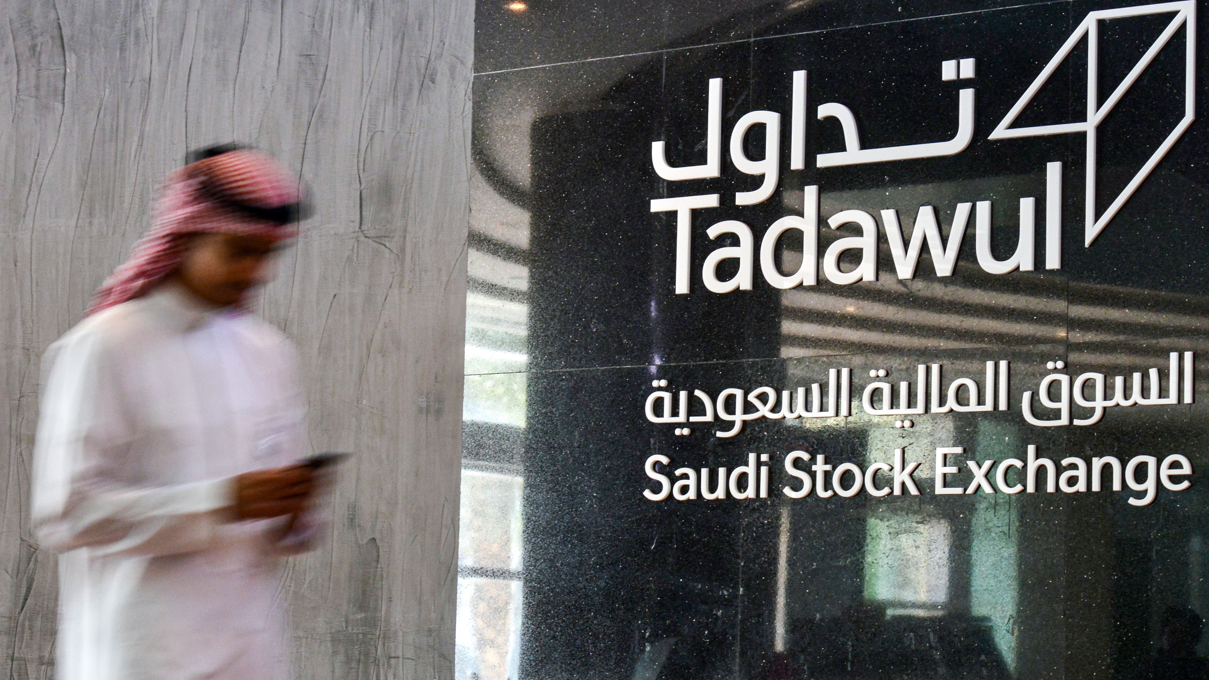 The Tadawul Saudi Stock Exchange logo on a dark background, with a person in traditional dress blurred in the foreground.