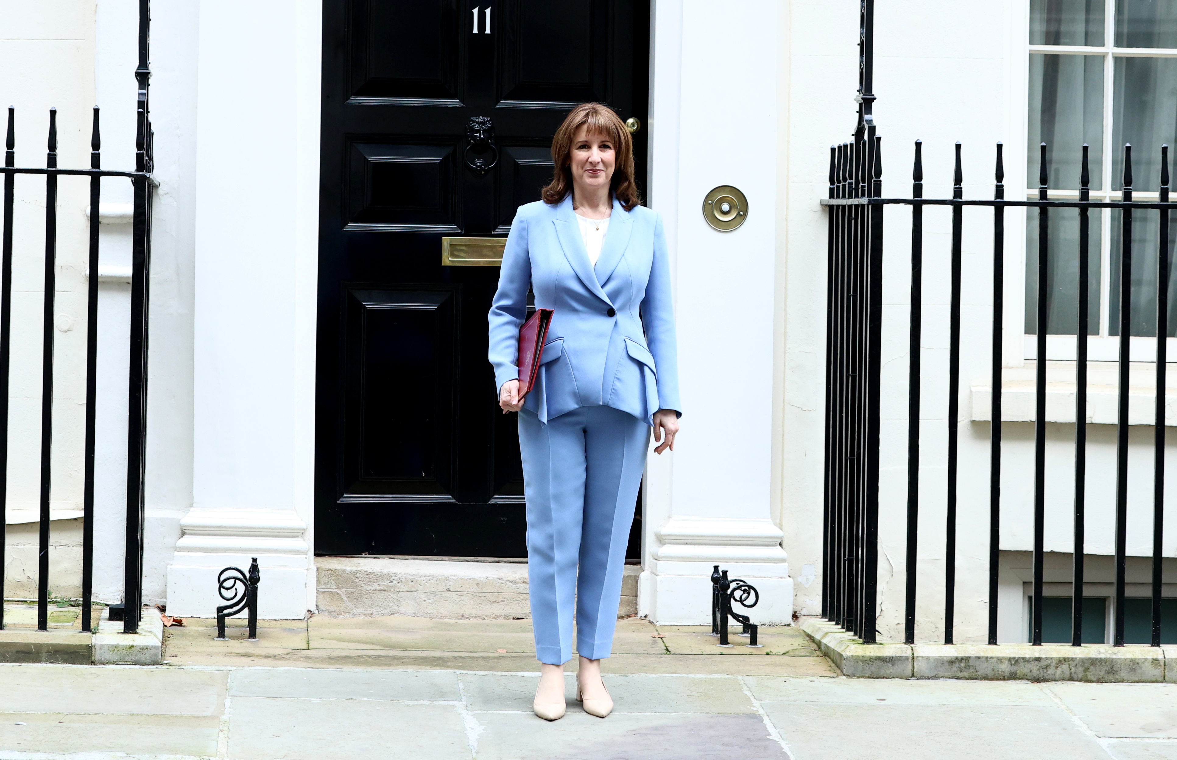 Rachel Reeves, Chancellor of the Exchequer, in a light blue pantsuit, stands in front of 11 Downing Street, holding a red folder.