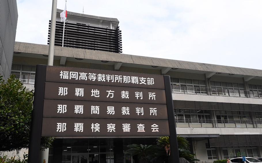 A modern multi‑story government building with a large vertical sign in front displaying Japanese text. The building has horizontal concrete lines, many windows, and a Japanese flag flying from a pole on the roof.