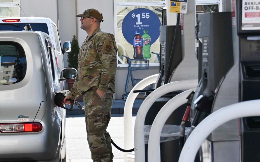 A service member in uniform stands at a gas pump, refueling a small silver car.