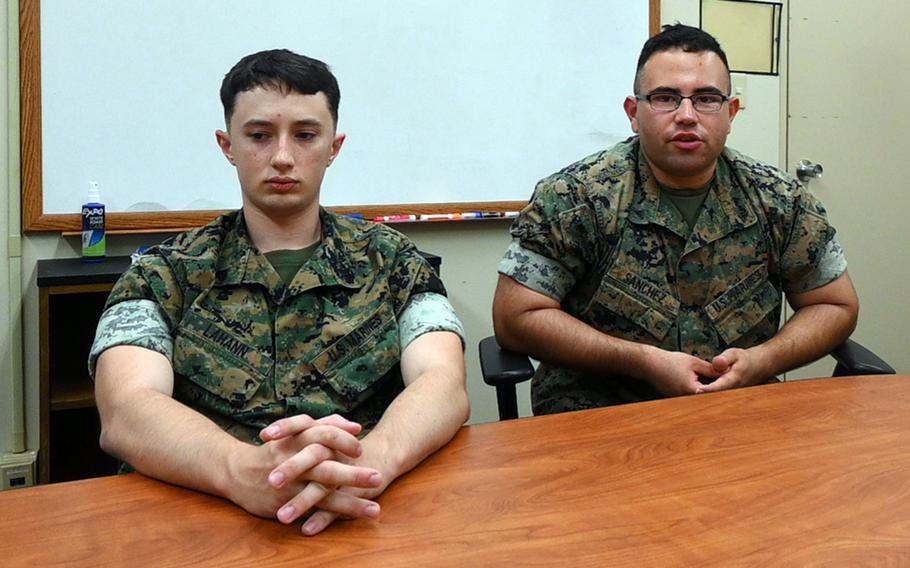 Two Marines in camouflage uniforms sit next to each other at a conference table, with a white dray erase board on the wall behind them.