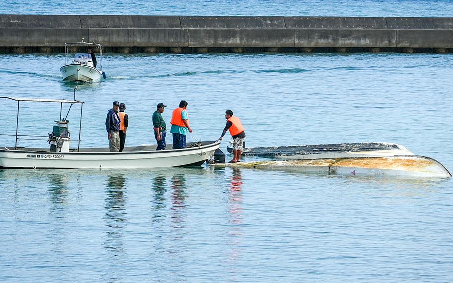 Four people, some of whom are wearing safety vests, stand on a boat next to a capsized boat as one stands on the hull of the capsized boat.