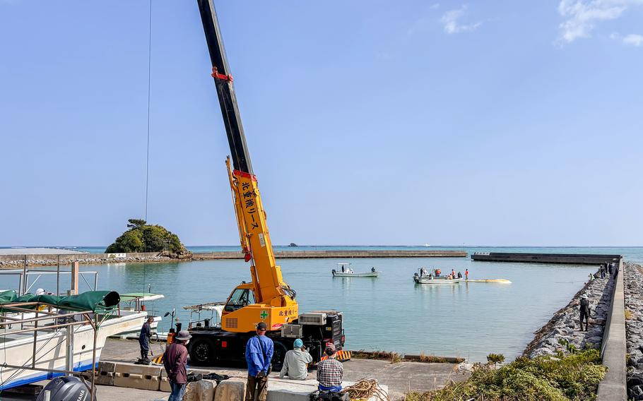 A crane sits in the foreground as two boats approach a capsized boat in the background. 