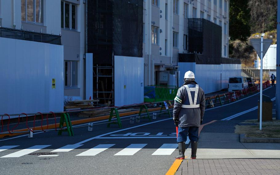 A construction worker in a blue reflective coat and white hard hat, seen from behind, stands on the side of a road, with green barriers blocking a lane next to a multi-story building.