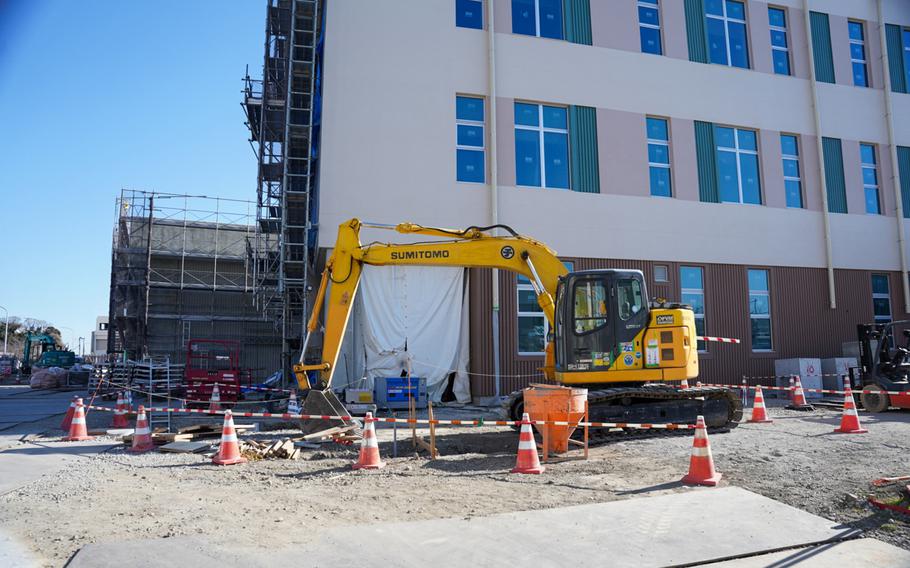 A yellow construction backhoe digs a hole in a patch of dirt in front of a multi-story building, with scaffolding on the left side and orange cones cordoning off the area inthe foreground.