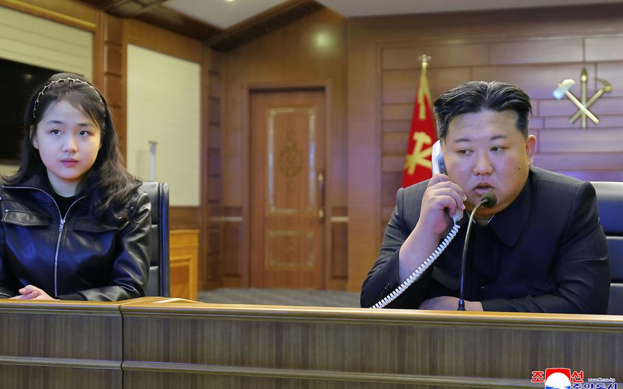 Kim Jong Un speaks on a telephone while seated at a desk with his daughter.