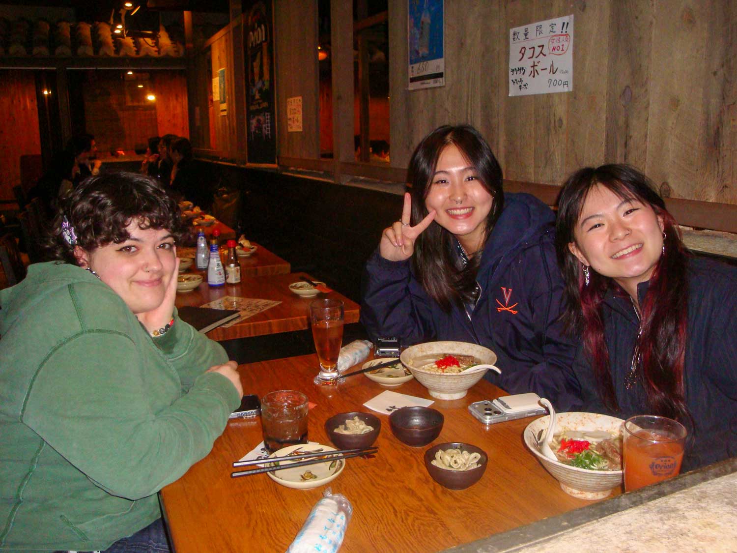 Three friends smiling at a restaurant table with bowls of ramen.
