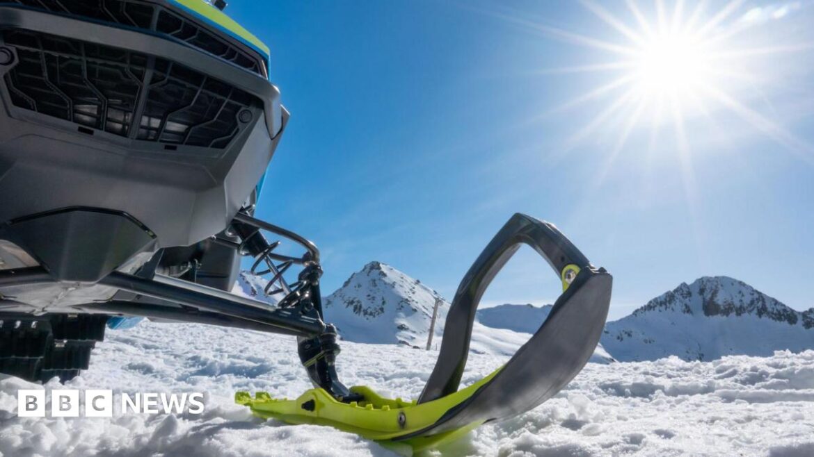 A close-up partial view of a snowmobile with snow and mountains in the background.