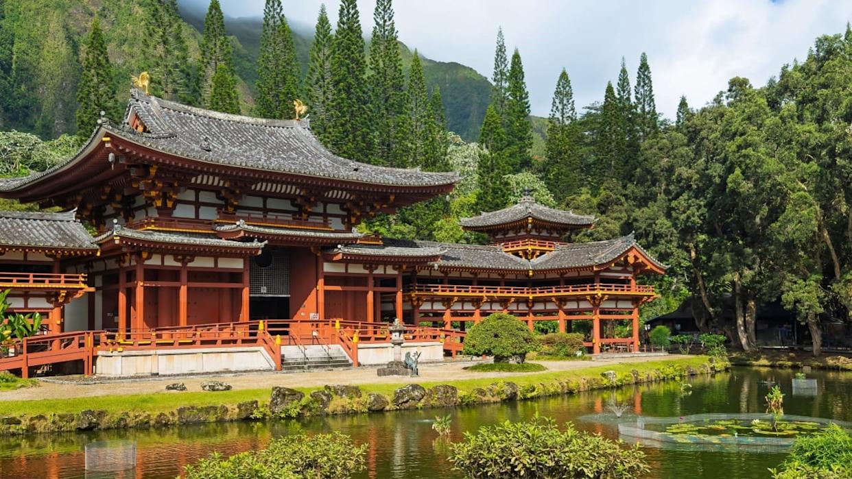 Kaneohe, Oahu, Hawaii, USA. - July 12, 2025: Maroon walls and gray roof, Byodo-In Buddhist temple front facade with koi-pond in front, green trees and mountain range in back under blue cloudscape