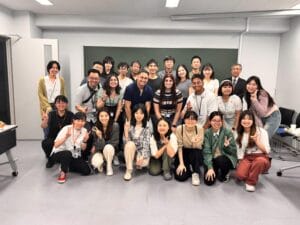 A group of people poses for a photo in a classroom, with a chalkboard in the background. Most are smiling and making peace signs.