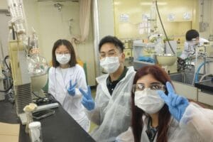 Three people wearing lab coats, gloves, and masks pose in a laboratory, making peace signs with their hands; lab equipment is visible on the tables.