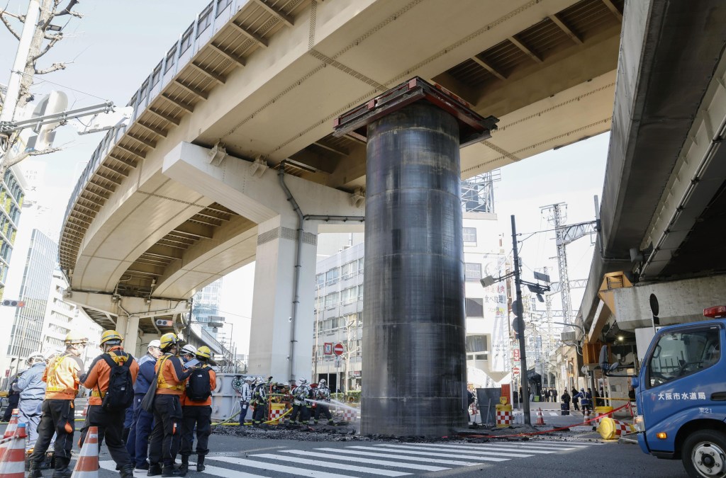 People in hard hats and vests gather around a large pipe that emerged from the ground at a sewer construction site under a highway overpass in Osaka, Japan.