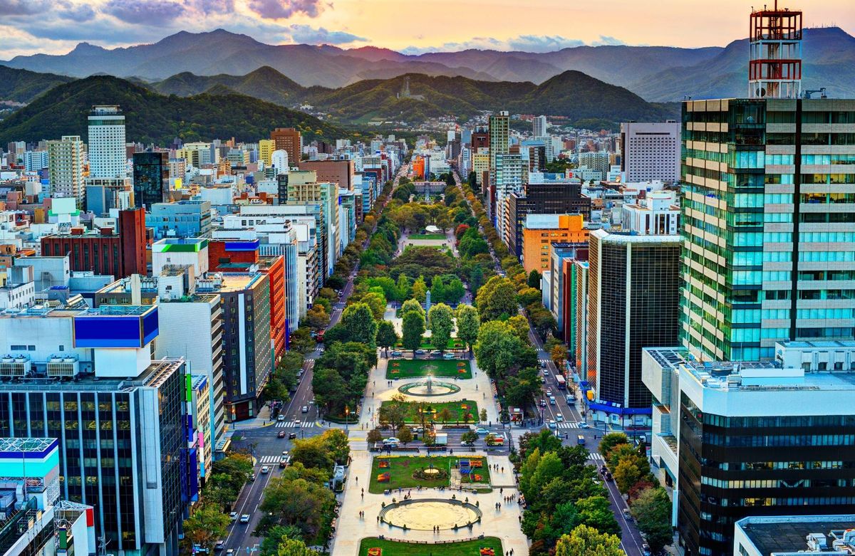 An aerial view of a bustling metropolitan area with high-rise buildings, a central park, and a backdrop of mountains under a setting sun.