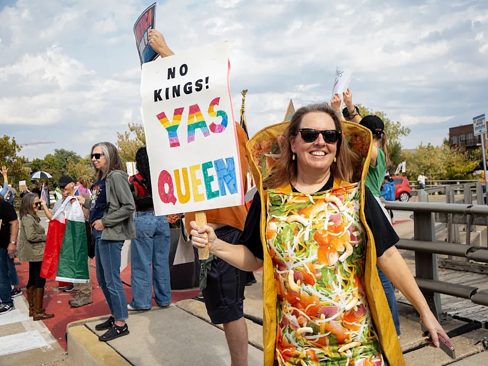 Person in a taco costume holding a sign saying "No Kings! Yas Queen" at a public protest. Other attendees hold signs in the background