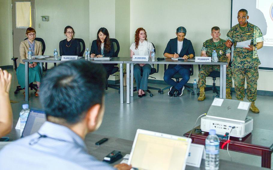 A Marine in uniform stands in front of a group of seated people and speaks into a microphone.