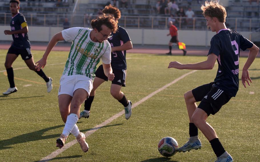 A high school soccer player in a black uniform dribbles the ball in front of a defender in a white uniform with green stripes.