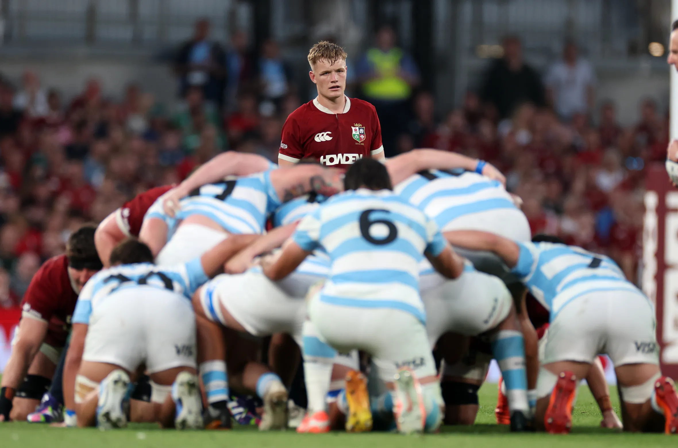 A rugby player in a red jersey, Tommy Freeman, stands looking forward as players in light blue and white jerseys are hunched over in a scrum.