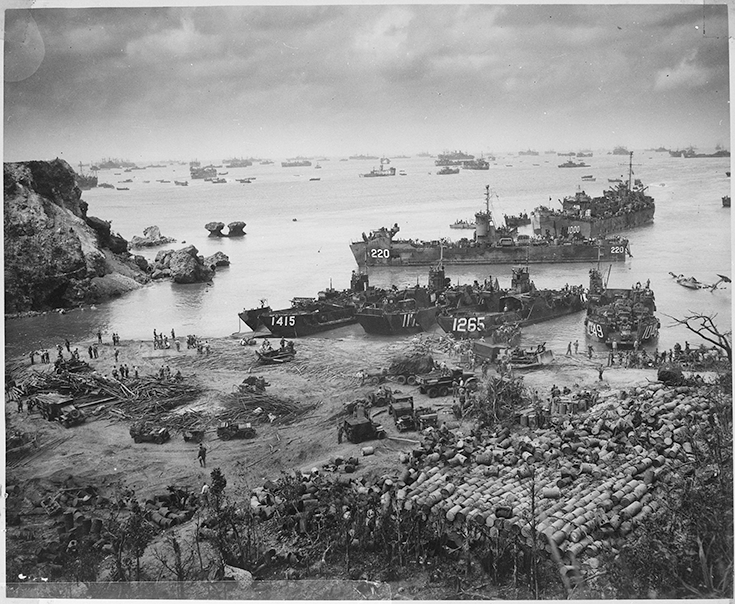 A horizontal photograph in black and white shows ships landing on a Japanese beach, with supplies and Soldiers on its shore. This image is the second in a series of six accompanying the article.