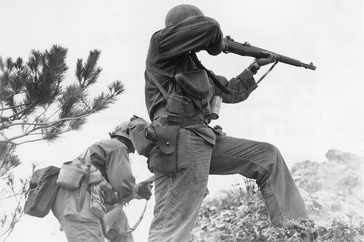 A horizontal photograph in black and white, shot from a low angle, shows a pair of helmeted Soldiers advancing among rocky foliage. One raises a rifle before him, a pistol holstered at his hip. His companion has a knife sheathed at his side, and he is raising or lowering his rifle. This image is the fourth in a series of six accompanying the article.