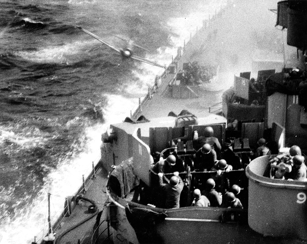 A square photograph in black and white shows an airplane flying toward the side of a battleship. Helmeted Soldiers can be seen on the ship’s deck, manning a gun mount. This image is the fifth in a series of six accompanying the article.