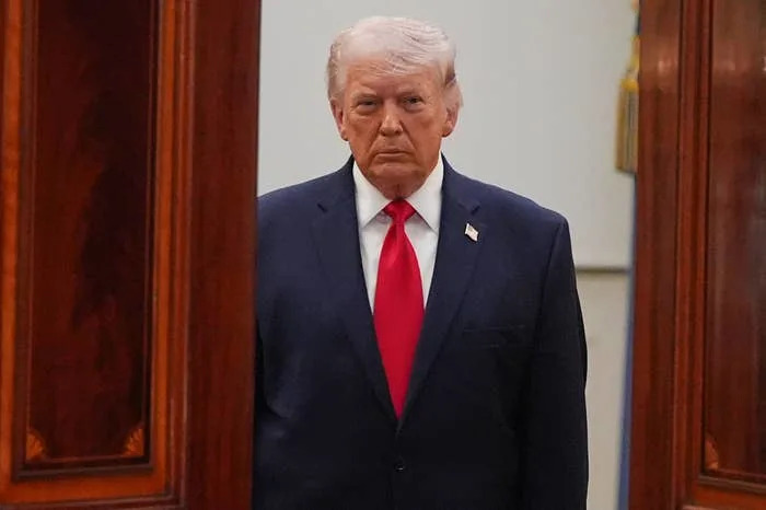 A person in a suit with a red tie stands between wooden panels, looking directly ahead
