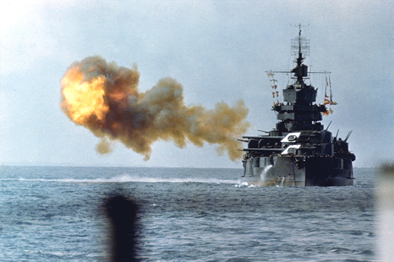 A horizontal photograph in color shows a battleship on the ocean, flames and smoke bursting in the blue sky beside it as it fires. This image is the sixth in a series of six accompanying the article.