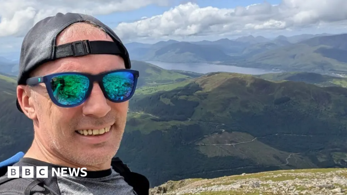 A man wearing green-tined sunglasses, a black cap on backwards and a grey T-shirt is standing on a mountain. In the background is a view of rolling hills and a reservoir.