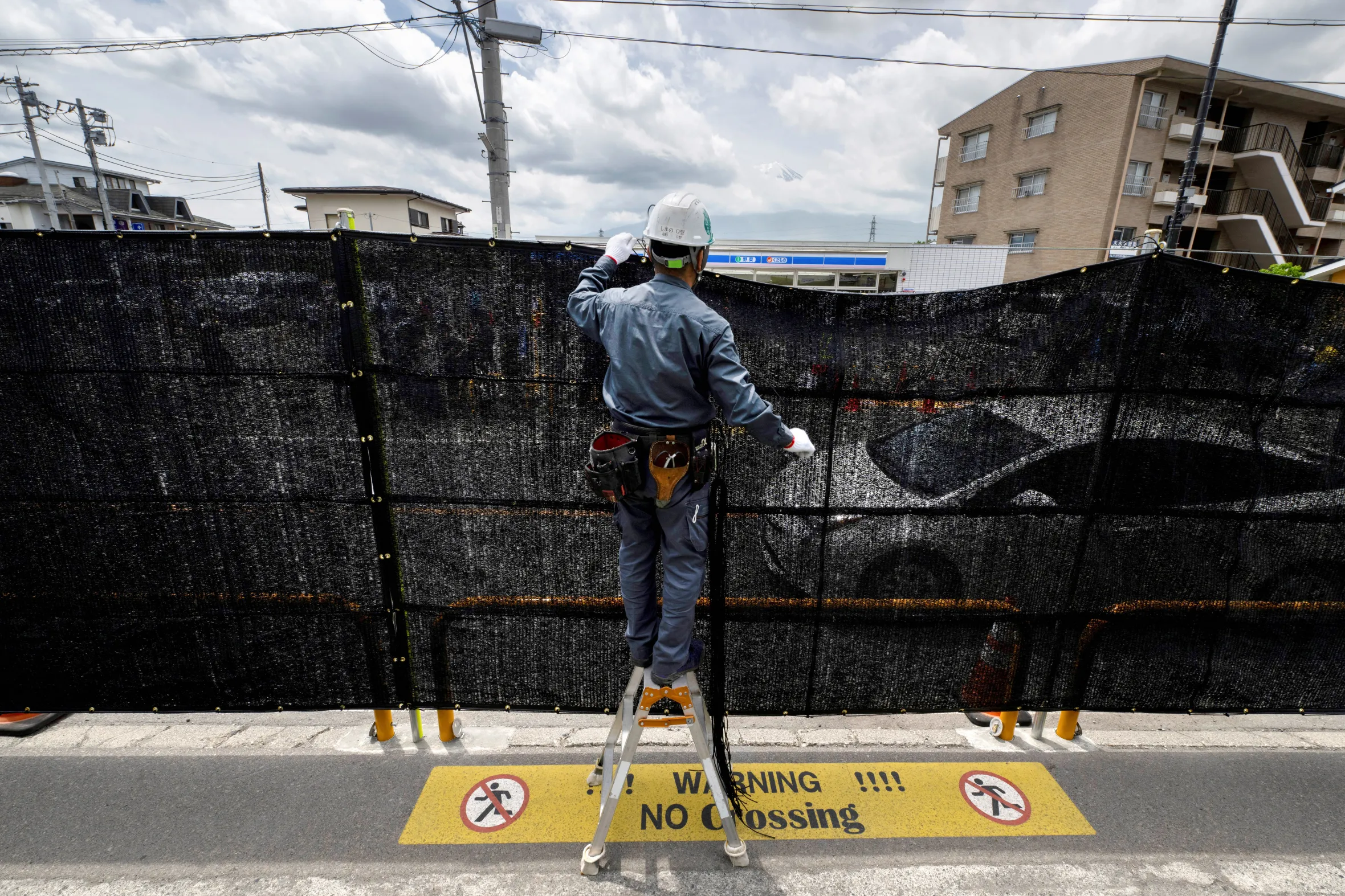 A worker installs a black mesh barrier to block the view of Mount Fuji, seen in the distance, from a convenience store.