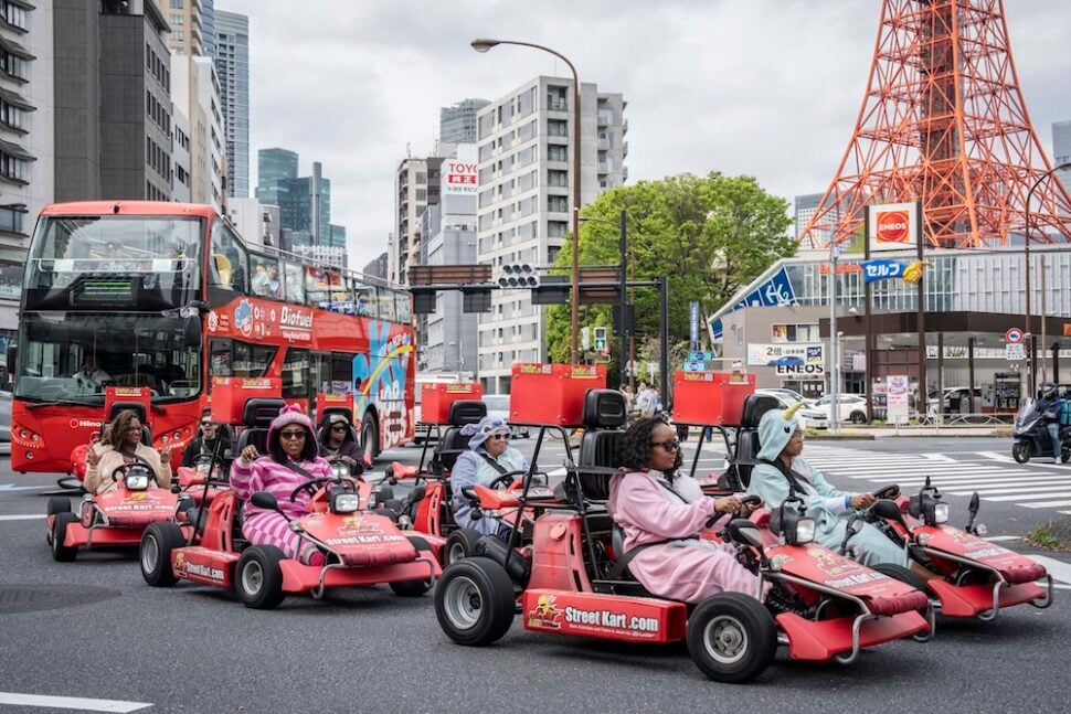 Group of tourists driving street go-karts through a Tokyo intersection with Tokyo Tower visible in the background.