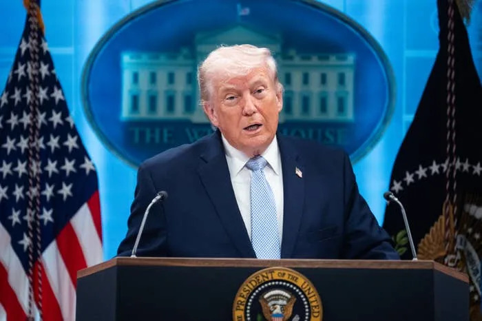 A person in a suit speaks at a podium with the U.S. presidential seal, in front of a backdrop featuring the White House