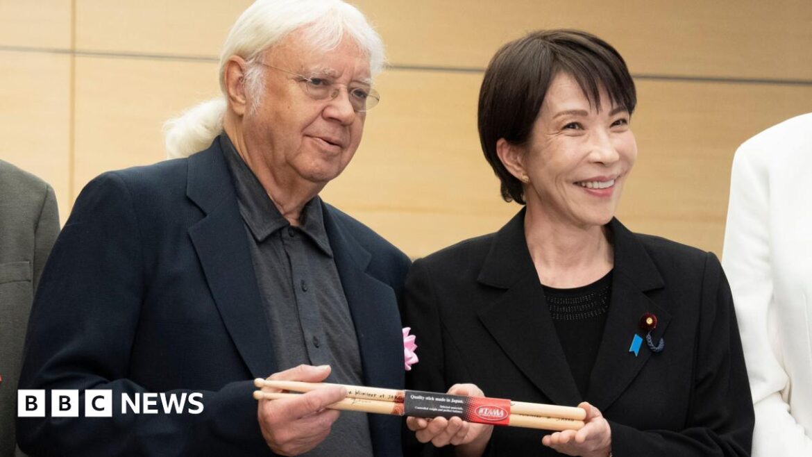 Japanese Prime Minister Sanae Takaichi poses with Ian Paice. They hold a set of signed drumsticks between them. Takaichi has short black hair and wears a black blazer over a black top. Paice has long white hair, tied back, and wears glasses, a black blazer and a grey buttoned shirt.