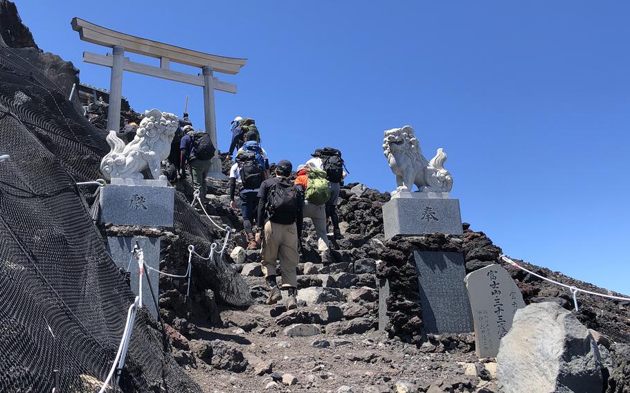 Hikers wearing backpacks and climbing gear, seen from behind and looking up, walk along a dirt trail up stone steps towards a wooden torii gate.