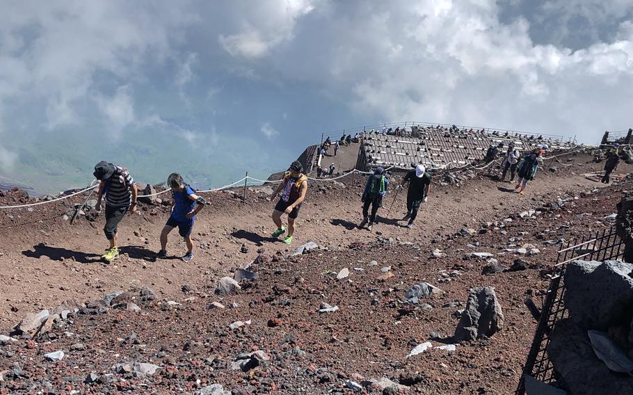 Hikers wearing backpacks and climbing gear, seen from a high angle, walk in a line along a dirt path, with swirling clouds behind them over the edge of a mountain.